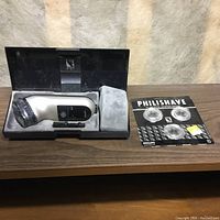 Philishave electric rotary razor model 905 in black and white, situated inside its open black case with a cleaning brush. To the right, a pack of replacement blades is displayed.