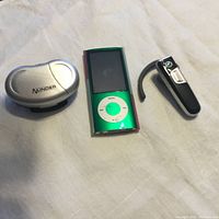 Photo of three items on a white fabric surface: green iPod Nano, black Bluetooth headset with ear hook, silver Minder walker pedometer.