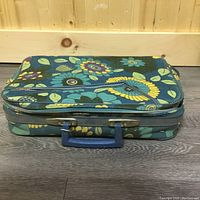 Vintage rectangular suitcase with colorful 1950s flower power cloth covering, blue zipper, and blue handle placed on wooden floor
