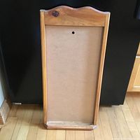 Wooden calendar holder frame resting upright against a black surface on a wooden floor, showing the back panel with hanging hole and natural wood finish.