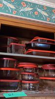 Various sizes of Anchor glass storage containers with red lids stacked in a wooden cabinet shelf.