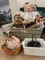 Plastic bin filled with multiple decorative baskets and plush Gund teddy bear on top, some baskets decorated with faux flowers.