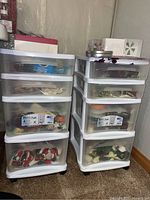 Two white plastic storage bins with four drawers each, filled with decorative crafting items such as cloth ornaments, stars, faux flowers, and thread.
