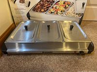 Front view of the Bravetti electric buffet server with three stainless steel chafing dishes with lids on, placed on carpet.