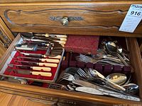 Open drawer revealing assorted stainless and silver plate flatware and boxed cutting utensil sets with cream handles.