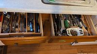 Open wooden drawers showing an assortment of stainless steel cutlery and metal kitchen tools including peelers and a jar opener.