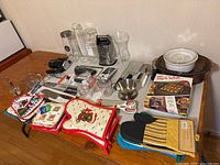 Photo showing various kitchenware items on wooden table including oven mitts, graters, glass canisters, ceramic dishes, spatulas, knives, and a cookbook.