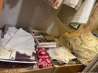 Photo showing stacked linens including doilies, folded cloths and packaged items with lace details and a patterned red/maroon item.