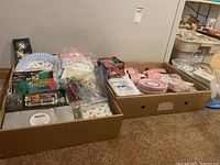 Overview of two boxes filled with assorted party supplies including paper plates, cutlery, stir sticks, cocktail forks, napkins, and cups.