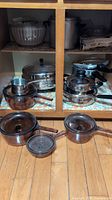 Wide view of the vintage glass pots with lids, stainless steel pressure cooker, and additional kitchen accessories inside a cabinet.