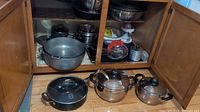 Cabinet with two Firenze stainless steel pots with lids, Lagostina frying pan with glass lid, and other kitchen utensils visible.