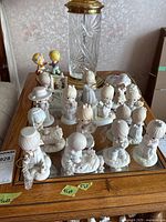All 14 Precious Moments porcelain figurines displayed on a mirrored surface table with the ceramic music box and heart-shaped Estee Lauder powder bowl nearby.