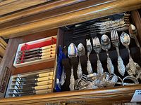 Drawer open displaying a set of Rogers flatware with ornate handles, including spoons and forks, and two boxed sets of cutting utensils with cream-colored handles.