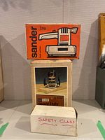 Stacked boxes showing Black & Decker sander box on top, Sears Craftsman power router box in the middle labeled 'Heavy Duty', and a white box labeled Safety Glass with glasses inside.