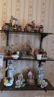 Three shelves showing two ceramic vintage cars on top shelf, multiple small cottage figurines and flower bouquets on middle shelf, and two standing figurines with flower bouquets on bottom shelf.