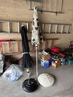 Two crank-lift patio umbrellas standing closed side by side in garage, with sand-filled umbrella stands, one black and one beige.