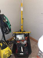 Pair of work lights with yellow metal frame and tripod, cords, trouble light, and lamp sockets inside black plastic bin, front view