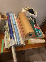 Photo showing multiple rolls of wrapping paper including several floral, metallic, and solid color rolls, a large brown packing paper roll, boxes of gift bags and tissue paper on a table.