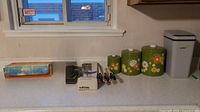 Photo displaying three vintage metal floral canisters in green with orange, white, yellow flowers and white lids, Wiltshire kitchen knives on stand, electric carving knife, motion detector, soil tester, and small grey-white plastic garbage can on white counter below window.