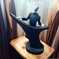 Full view of black and silver Art Deco style sculpture showing man playing a grand piano on a rounded base, placed on a wooden surface with curtain background.