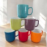 Set of six colorful ceramic mugs arranged in a pyramid stack on a wooden surface with daylight illumination. Visible vibrant colors blue, red, orange, lime green, purple, aqua. White interior on all mugs.