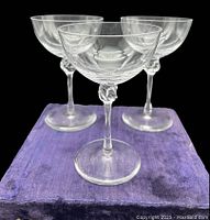 Three clear crystal footed champagne glasses with unique twisted hand-like design on the stem, placed on a purple cloth against a black background.
