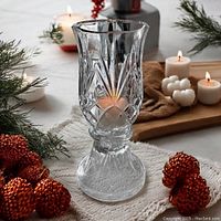 Clear crystal candle lamp with diamond pattern lit by a candle inside, placed on a cloth surface with decorative pine branches and round ornaments nearby.
