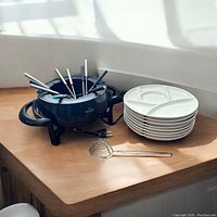 Electric fondue pot, six color-coded skewers, eight white sectioned plates stacked, and a metal scoop on wooden surface near window with sunlight.