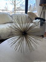 Full view of gold-coloured spiked ball metallic decorative piece, spherical with numerous thin spikes radiating outward, displayed indoors on a table near a window.