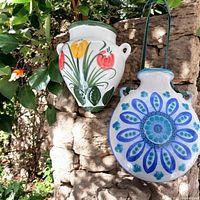 Two ceramic wall pockets with floral hand painted designs hanging outdoors on a textured stone wall among green foliage, showing the white tulip design piece with handles on the left and the blue circular floral design on the right with velvet ribbon.