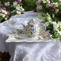 Ceramic teapot and serving tray placed on a white lace tablecloth on an outdoor table with flowers in the background.