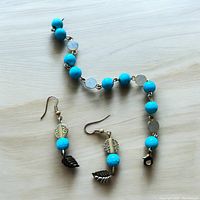 Pendant earrings with turquoise colored beads, gold-tone accents, and dark leaf charms next to a bracelet of matching turquoise and translucent beads chained together with gold-tone links, on a pale wood surface.
