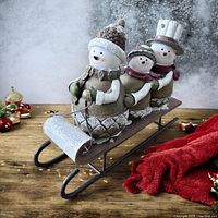 Snowmen friends sledding tabletop decor shown from side angle on wooden surface with festive holiday items in the background.