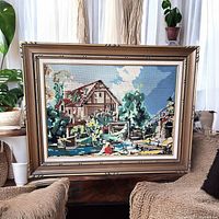 Frontal view of the framed needlepoint artwork showing the full detailed grist mill summer scene with woman by the stream, vibrant colors and solid wood frame.