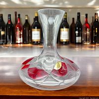 Clear glass mosaic decanter with red and gold accents shown on a wooden surface with bottles in the background.