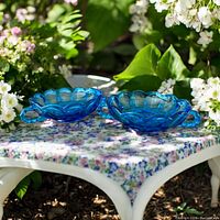 Pair of colonial blue Fenton glass candy/nut dishes with scalloped edges and looped handles shown on floral table outside.