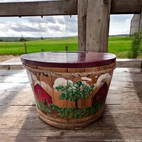 Side view showing the handpainted country scene with red barns and trees on the wooden round crate sitting on rustic wood surface outdoors.