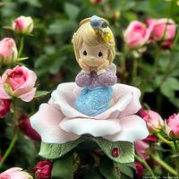 Front angled view of the pink rose figurine with the girl sitting inside the petals, surrounded by real pink rose flowers in the background.