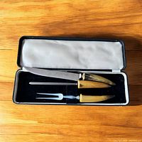 Set of three carving tools in an open black hinged case with satin lining inside the lid and velvet base, showing the knife, sharpening steel, and carving fork with matching horn-colored handles.