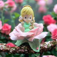 Figurine of a girl with blonde hair sitting inside a pink rose on a branch with blurred pink flowers background.