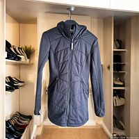 Front view of black mid-length zip-up hooded jacket with geometric stitched panel detailing, hanging on black hanger, showing zipper and hood.