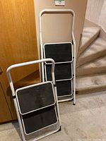 Two white metal frame step ladders with two black steps each, shown unfolded and leaning against a wall near stairs.