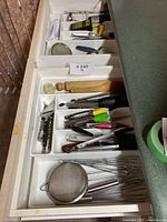 Top view of two kitchen drawers holding various cooking utensils including mesh strainer, whisks, tongs with colored silicone tips, peelers, measuring spoons and brushes.