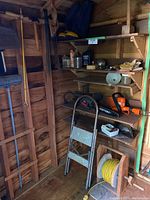 View of wooden shed interior showing gardening tools hanging on wall, metal step ladder, yellow extension cord on spool, and shelves with power tools and items.