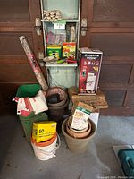 Overview photo showing assorted gardening pots, boxes of slug pellets, paper bags, gloves, and gardening tools stacked in front of garage door.