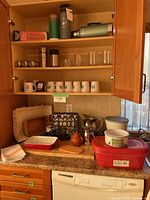 Kitchen cabinet with upper shelf showing green Stanley thermos, glass mugs, and blue Trading Post Brewing bottle, vintage thermos, plus lower counter showing Pyrex dishes, coffee pot, wooden trays, and kitchen linens.