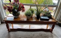 Long narrow polished wooden coffee table with decorative carved edges and legs, placed in front of a window with plants on top.