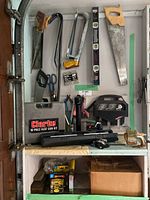 Clarke heat gun kit case and various hand tools including saws, clamps, levels, and a black handled saw frame on a shelf with boxes below.