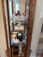 Display cabinet showing glass decanters under glass domes, bone china items, and crystal shot glasses arranged on shelves.