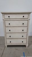 Front view of the white washed five drawer chest showing all drawers and knobs.